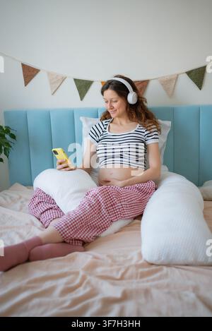 Donna incinta che ascolta musica con le cuffie, si connette con il bambino non ancora nato, pratica la meditazione per il parto per il benessere materno e il rilassamento durante la maternità Foto Stock