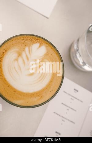 Vista dall'alto di un latte con decorazione in schiuma a forma di foglia accanto a un bicchiere d'acqua e ai menu del caffè Foto Stock