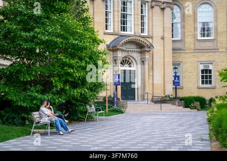 Toronto, Ontario, Canada - 06 21 2025: Una giovane coppia o studenti che si rilassano e conversano su una panchina fuori da un classico edificio accademico in arenaria Foto Stock