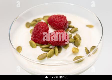 Una foto ravvicinata di cibo che mostra uno yogurt greco cremoso servito in una ciotola di vetro trasparente, ricoperto da una generosa spolverata di semi di zucca nutrienti e guarnito Foto Stock