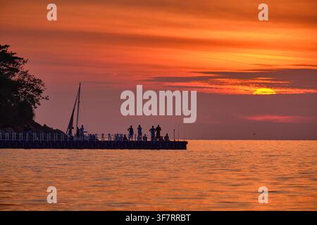 Koh Lipe, Thailandia - 20 gen. 2025: Silhouette di persone su un molo di legno che osservano un vivace tramonto arancione con il grande sole sopra l'oceano calmo a Sunset Beach. Foto Stock