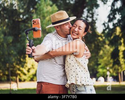 Giovane coppia allegra che si abbraccia e ride mentre si filma con uno stabilizzatore per smartphone all'aperto. Foto Stock