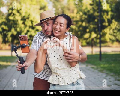 Giovane coppia felice che si abbraccia calorosamente mentre crea contenuti per i social media con uno smartphone all'aperto. Foto Stock