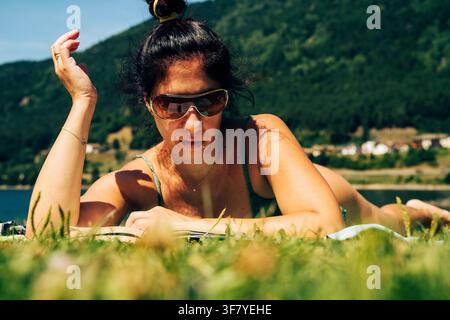 Una donna legge un libro mentre è sdraiata su un prato vicino alla riva del Lago di Piazze in Trentino. La donna ha lunghi capelli scuri e occhiali da sole sul viso Foto Stock