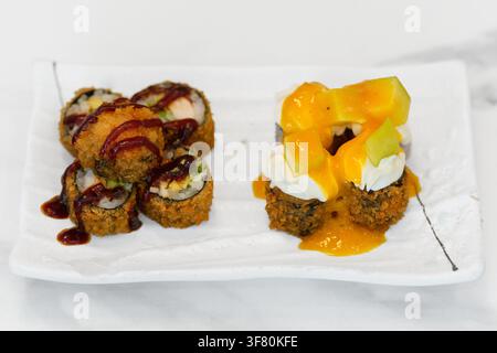 Vista ad angolo alto di involtini di sushi tempura fritti serviti su un piatto di ceramica bianca. Metà è ricoperta da salsa unagi scura e l'altra metà è decorata con formaggio cremoso, Foto Stock