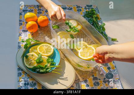 Mani che preparano una bevanda rinfrescante agli agrumi con limoni a fette, arance e menta fresca alla luce naturale brillante. Foto Stock