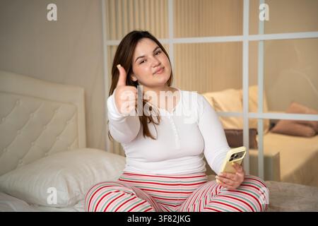 Giovane donna sorridente seduta sul letto che dà il pollice in su mentre tiene uno smartphone. Comunicazione mobile di feedback positivo e gesto di approvazione del cliente. Foto Stock
