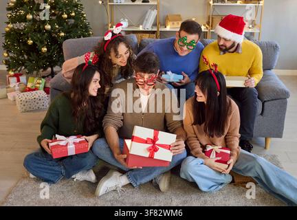Gruppo di giovani amici che scartano regali di Natale a casa ridendo con cappelli festivi Foto Stock