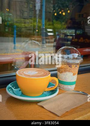 Tazza gialla di latte fresco con latte art a forma di cuore accanto a un dessert cremoso al chia su un tavolo di legno del caffè, riflessi nel vetro della finestra. Caffè luminoso Foto Stock
