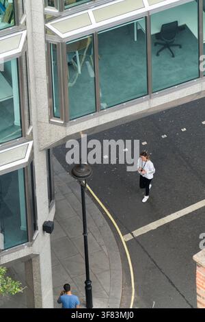 Una donna corre davanti a un moderno edificio per uffici nel distretto finanziario di Londra, probabilmente in viaggio verso il lavoro o in pausa nel mezzo della frenetica giornata cittadina. Foto Stock