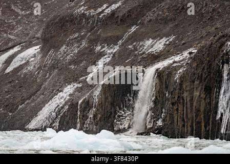 Cascata scorre lungo un pendio roccioso verso acque ghiacciate mentre una coltre invernale copre le scogliere in Groenlandia Foto Stock