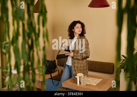 Donna dai capelli ricci lavora da remoto sul suo laptop godendosi un'atmosfera accogliente da caffè Foto Stock
