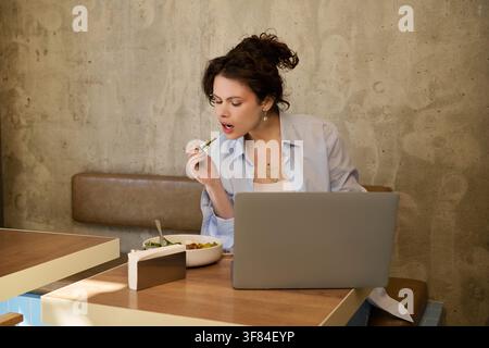 Donna dai capelli ricci si impegna nel lavoro da remoto sul suo laptop mentre assapora il suo pasto Foto Stock