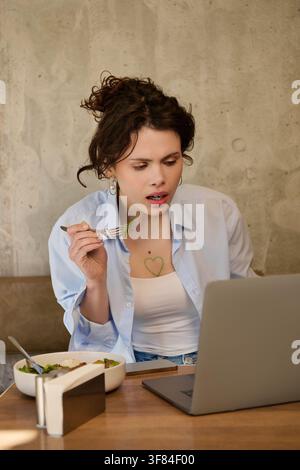 Giovane donna dai capelli ricci interagisce con il suo laptop a un tavolo di caffè vivace mentre mangia insalata Foto Stock