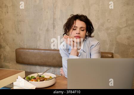Donna dai capelli ricci si impegna con il suo laptop a un tavolo accogliente del caffè mentre mangia Foto Stock