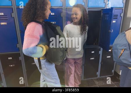 Giovani donne che chiacchierano nel corridoio della scuola accanto agli armadietti blu, indossano un maglione pastello e un maglione color crema. Corridoio, studenti, zaini, a righe, lavorato a maglia, c Foto Stock