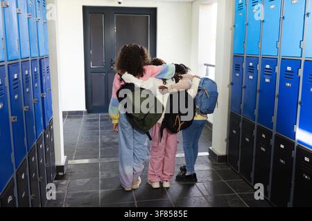 Tre adolescenti camminano nel corridoio della scuola in una maglietta pastello portando zaini davanti agli armadietti sotto una telecamera. Ragazze, amicizia, corridoio, colorato, casual, snea Foto Stock