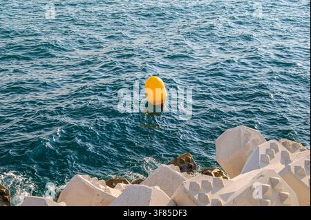 Una boa giallo brillante galleggia su acque blu agitate vicino a un frangiflutti di cemento. Foto Stock