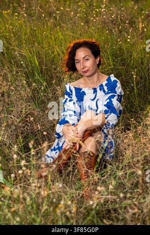 Donna sorridente seduta in un prato illuminato dal sole, indossa un vestito blu a spalle scoperte e stivali con lacci marroni, ritratto dello stile di vita rilassato. Foto Stock