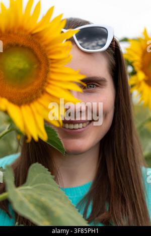 Donna che tiene un girasole, in un campo di girasoli, con girasole, occhiali da sole, da una prospettiva in primo piano, con accenti gialli, verdi e un'atmosfera felice Foto Stock