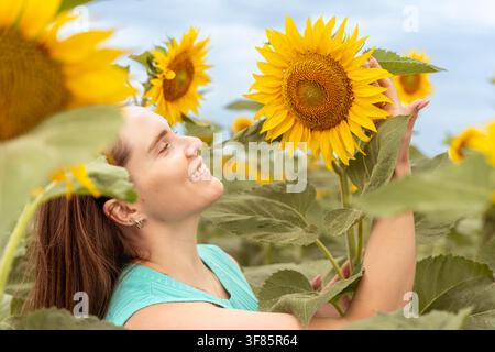 Donna che tiene un girasole e sorride in un campo di foglie verdi, vista laterale primo piano, moda viaggio naturale, giallo brillante e verde Foto Stock