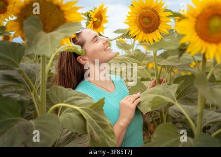 Donna che sorride e guarda i girasoli, in un campo di girasoli, con girasoli, abbigliamento femminile, da una prospettiva a livello degli occhi in primo piano Foto Stock