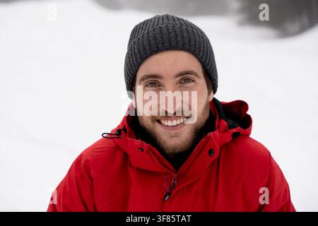 Ritratto ravvicinato di un giovane allegro che indossa una giacca rossa e un cappello beanie grigio, guardando direttamente la fotocamera Foto Stock