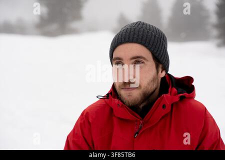 Giovane uomo con giacca rossa e berretto in piedi in un paesaggio invernale innevato, che guarda lontano con un'espressione contemplativa Foto Stock