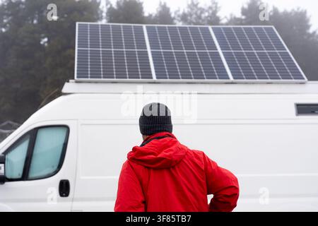 Veduta posteriore di un uomo con una giacca rossa in piedi accanto al suo camper bianco con pannelli solari fotovoltaici sul tetto, che rappresenta l'energia pulita Foto Stock