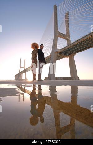Coppia alla moda in piedi schiena contro schiena vicino a una pozzanghera che riflette il ponte Vasco da Gama al tramonto Foto Stock
