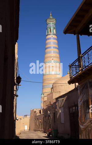 Minareto di Islam Khoja visto attraverso un vicolo stretto a Ichan Kala, Khiva. Bande di piastrelle a righe e cupola verde si ergono sopra i muri di mattoni di fango sotto un cielo blu terso. Ideale per guide turistiche e l’Asia centrale. Foto Stock