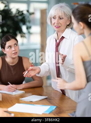 Dipendente d'ufficio matura concentrata in piedi con una penna in mano vicino a un tavolo dove colleghi seduti discutono emotivamente problemi Foto Stock