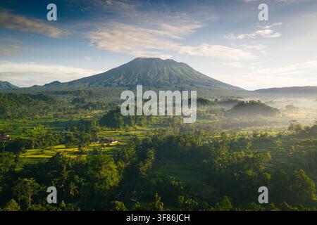 Vista aerea del Monte Agung che si erge sopra le rigogliose terrazze di riso verdi e la valle nebbiosa di Sidemen, Bali, Indonesia Foto Stock