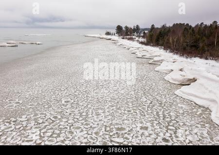 Vista aerea del faro di Sturgeon Point circondato da banchi di ghiaccio sulla costa ghiacciata del Lago Huron con alberi innevati sotto un cielo nuvoloso a H Foto Stock