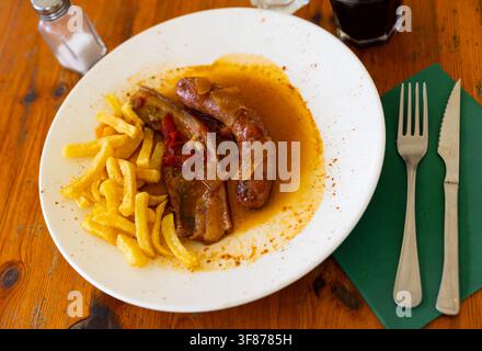 Gustose salsicce e pancetta arrosto in salsa di pomodoro insieme a patatine fritte servite su un tavolo di legno in un caffè Foto Stock