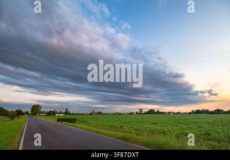 Strada di campagna che attraversa campi verdi sotto un cielo drammatico al crepuscolo dopo il passaggio di un temporale sulla Val Padana, Italia. Foto Stock