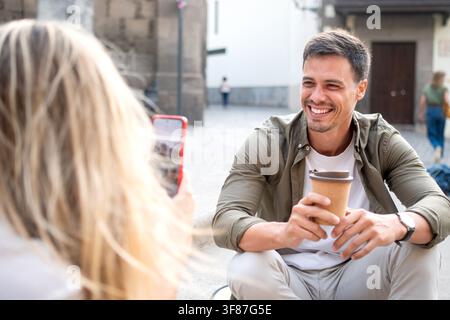 Donna fotografa il suo fidanzato sorridente che tiene un caffè da asporto. Foto Stock