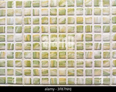 Una parete presenta piccole piastrelle verdi e bianche disposte in un motivo a griglia con una texture ruvida, aggiungendo profondità e interesse alla superficie. Foto Stock