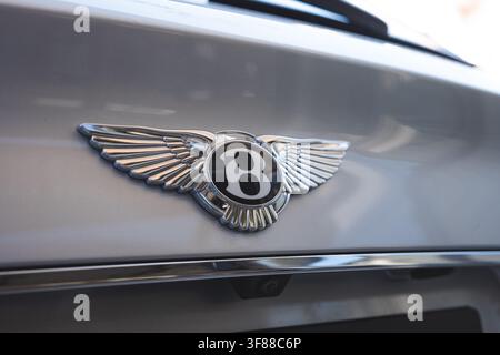 Lindau, Svizzera, 9 agosto 2026. Dettagliato primo piano di un badge alare cromato Bentley con emblema della lettera B su un cofano metallico chiaro. Foto Stock