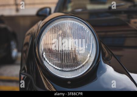 Scatto ravvicinato dettagliato di un modulo faro rotondo vintage in vetro H4 integrato nel pannello paraurti anteriore nero lucido di un'auto sportiva classica Porsche 964. Foto Stock