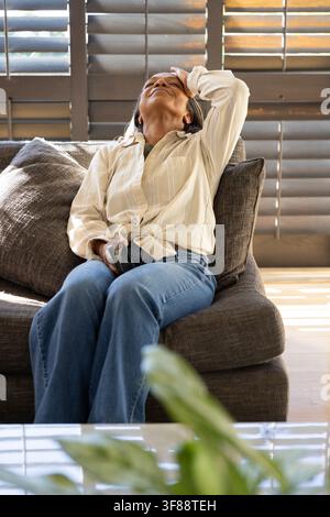 Donna africana matura seduta su un divano grigio scuro in salotto, tiene uno smartphone, tocca la fronte. Luce solare, ombre, persiane, divano, cuscino, pianta Foto Stock