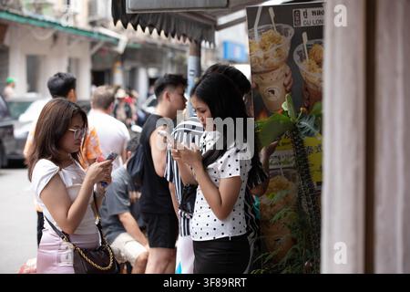 BANGKOK, THAILANDIA - 8 AGOSTO 2026: Turiste femminili che utilizzano uno smartphone mentre sono in fila per il cibo, profilo laterale, strada Song Wat. Foto Stock