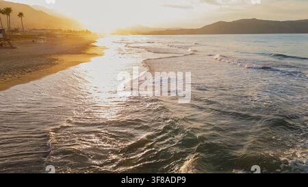 Vista aerea con drone di dolci onde del mare che lambiscono una riva sabbiosa al tramonto con costa di palme e silhouette di montagne nella calda luce dorata del sole, foto stock Foto Stock