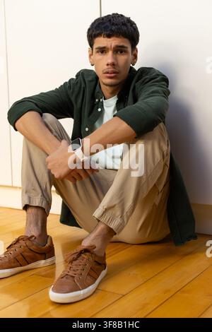 Uomo afroamericano seduto sul pavimento in legno duro, appoggiato a mobili bianchi, mostra uno smartwatch. Ritratto, lifestyle, minimal, interno, caldo, casual Foto Stock