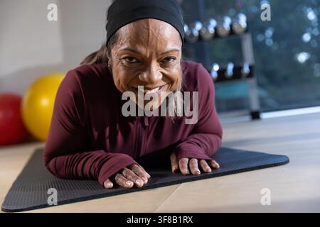 Donna matura che esegue planche sugli avambracci su un tappeto nero in palestra con manubri, indossa un top bordeaux. Fitness, studio, addominali, stabilità, pesi, naturale, lig Foto Stock