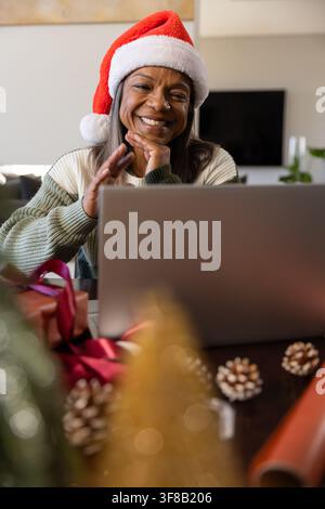 Donna di mezza età con cappello di Babbo Natale in maglione seduta a casa usando un laptop, regali incartati, pigne. Accogliente, festoso, Natale, soggiorno, lavoro da casa Foto Stock