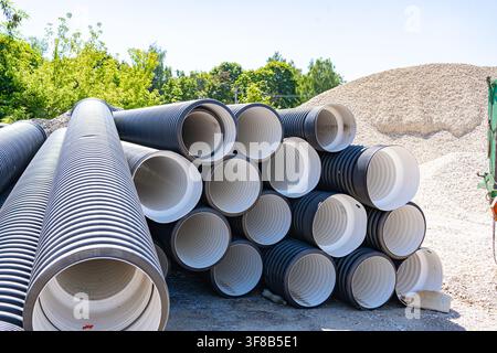Pila di grandi tubi di plastica ondulata nera conservati all'aperto vicino a cumuli di ghiaia in un cantiere edile. Ideali per impianti idraulici comunali, sistemi di drenaggio Foto Stock