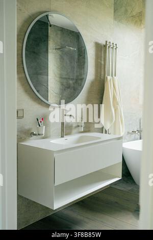 Design contemporaneo di bagno con mensola sospesa bianca per lavabo, specchio rotondo e rail riscaldante per asciugamani. Perfetto per architettura di lusso, idraulica Foto Stock