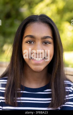 Adolescente afroamericana sorridente seduta all'aperto su una panchina, indossa un top a righe blu navy. Ritratto, felicità, natura, luce del sole, bokeh, casual Foto Stock