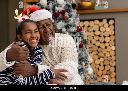 Nonno africano e nipote adolescente che si abbracciano sul divano a casa con albero di Natale, cappello di Babbo Natale. Accogliente, festoso, famiglia, calore, multigenerazionale Foto Stock